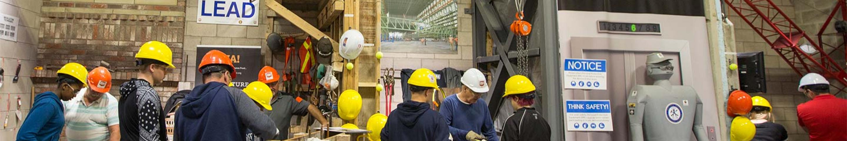 A mixture of youth and men working in a workshop while wearing yellow hard hats and gloves